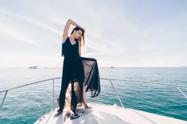 Una bella ragazza di lusso in un abito estivo nero in posa sul suo yacht bianco godendo di un viaggio in mare — Foto Stock