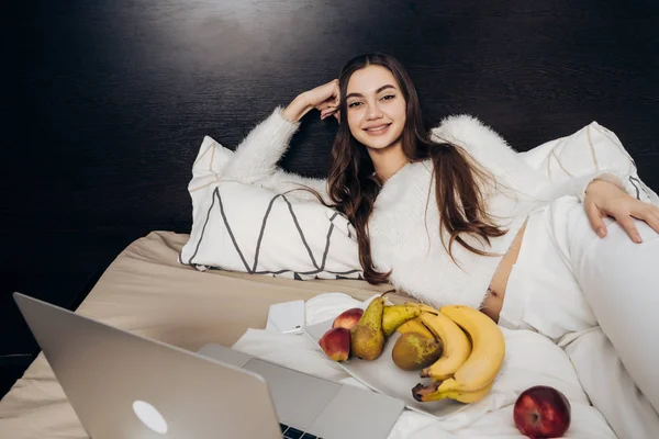Boldog hosszú hajú lány az ágyban, egy film a laptop, és eszik egészséges gyümölcs pihenő — Stock Fotó
