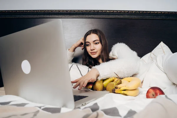 Мила дівчина відпочиває в ліжку після важкого робочого дня, дивиться цікавий фільм на ноутбуці і їсть фрукти — стокове фото