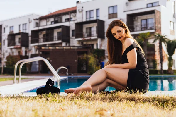 Uma mulher sexy de luxo em um vestido preto elegante descansando à beira da piscina ao sol, apreciando as vidas ricas ... — Fotografia de Stock