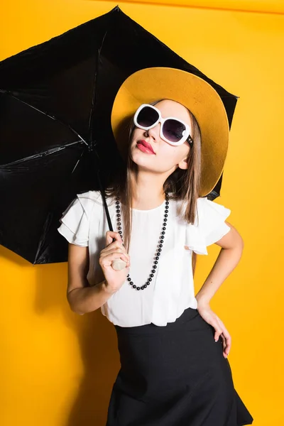 Menina elegante atraente em óculos de sol e chapéu guarda guarda-chuva e posando no fundo amarelo — Fotografia de Stock