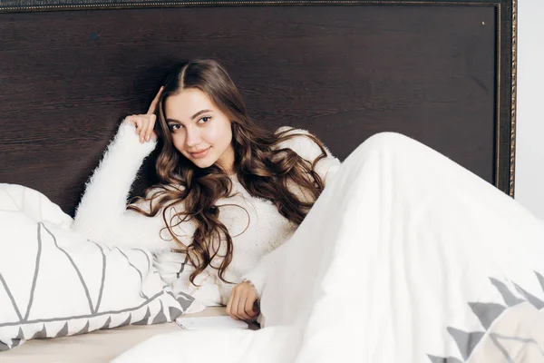 Encantadora chica de pelo largo en pijama blanco se encuentra en la cama, descansando después de un día de trabajo y sonriendo — Foto de Stock