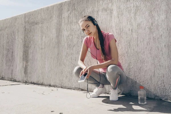 sports young girl resting after a hard workout outdoors, listening to music on headphones