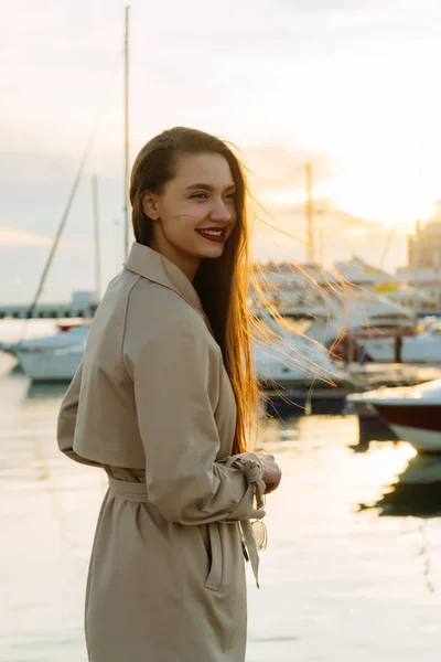 Luxuosa menina de cabelos longos feliz em um casaco bege posando junto ao mar ao pôr do sol — Fotografia de Stock