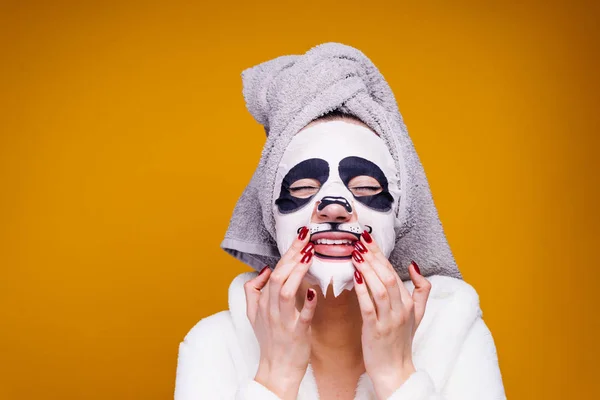 A smiling young girl with a towel on her head after a shower puts a useful moisturizing mask with a panda face on her face — Stockfoto