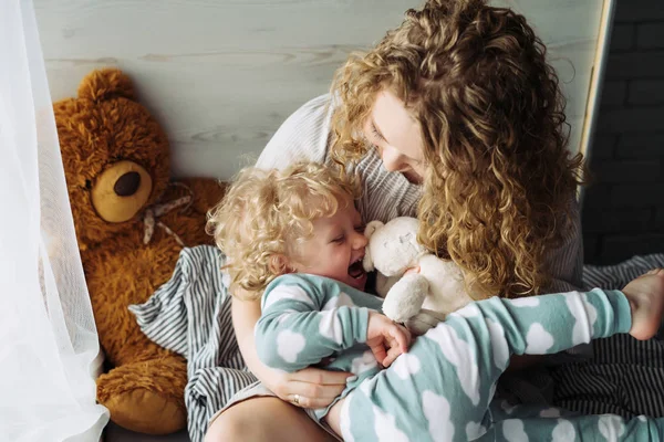 Caring curly mother plays with her little son, in blue pajamas, have fun and laugh — Stock Photo, Image