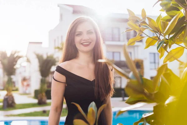 Lussuosa ragazza dai capelli lunghi in un abito nero in posa presso la piscina blu al sole, godendo di una vita ricca — Foto Stock