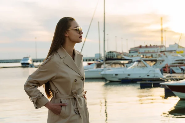 Confiada chica de lujo en gafas de sol y un abrigo beige está esperando su barco en el puerto al atardecer — Foto de Stock