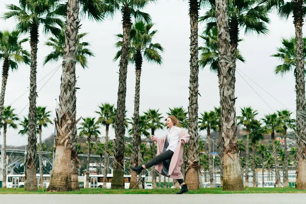 uzun zaman pembe bir ceket komik bir kadın parkta yanındaki uzun boylu yeşil palms yürür, baharda sevinir