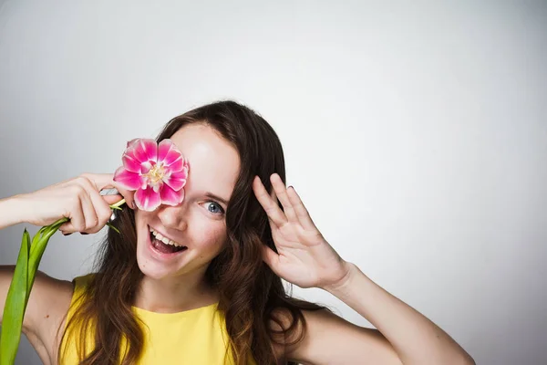 Divertida mujer de ojos azules en un vestido amarillo se ríe, sostiene una flor rosa fragante en la cara — Foto de Stock