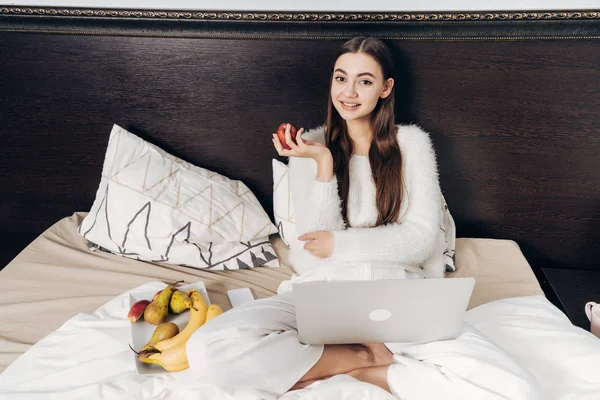 Lächelnde junge Frau wacht früh auf, sitzt im Bett und frühstückt mit nützlichem Obst — Stockfoto