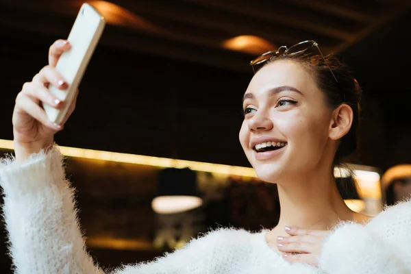 Szczęśliwy uśmiechający się dziewczyna w białym swetrze siedzi w przytulnej restauracji i sprawia, że selfie w swoim smartfonie — Zdjęcie stockowe