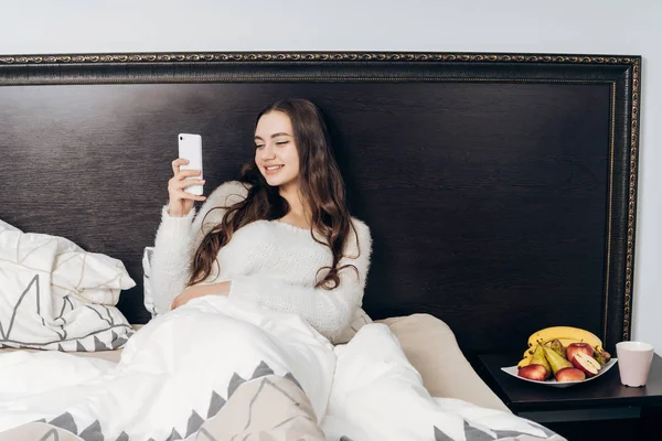 Sonriente jovencita en pijama se sienta en la cama por la noche, mira en su teléfono inteligente, quiere dormir — Foto de Stock