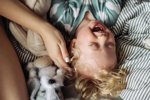 Pequeño lindo bebé niño en pijama azul acostado en la cama al lado de mamá y riendo —  Fotos de Stock