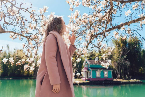 pembe bir ceket şık kadın bir su birikintisi yanındaki bir parkta güzel kokulu bir manolya kokuyor