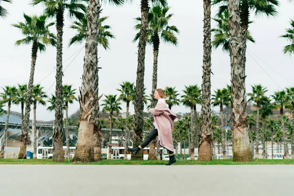uzun bir ceket garip bir şık kız sıcak uzun boylu yeşil palms, yanındaki bir parkta yürüyor