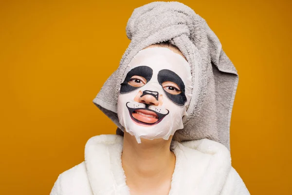 Смешная молодая девушка с полотенцем на голове улыбается, на лице увлажняющая маска с лицом панды — стоковое фото