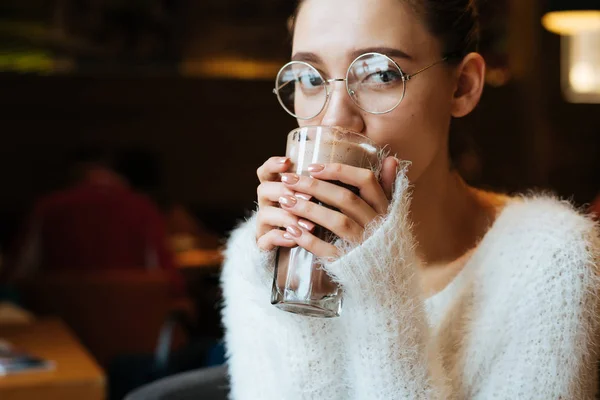 Молода дівчина студент в окулярах п'є гарячий латте в кафе після школи, дивиться на камеру — стокове фото