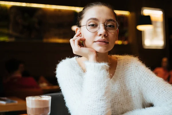 Hermosa joven con una chaqueta blanca y gafas está sentado en un acogedor café y beber café aromático —  Fotos de Stock