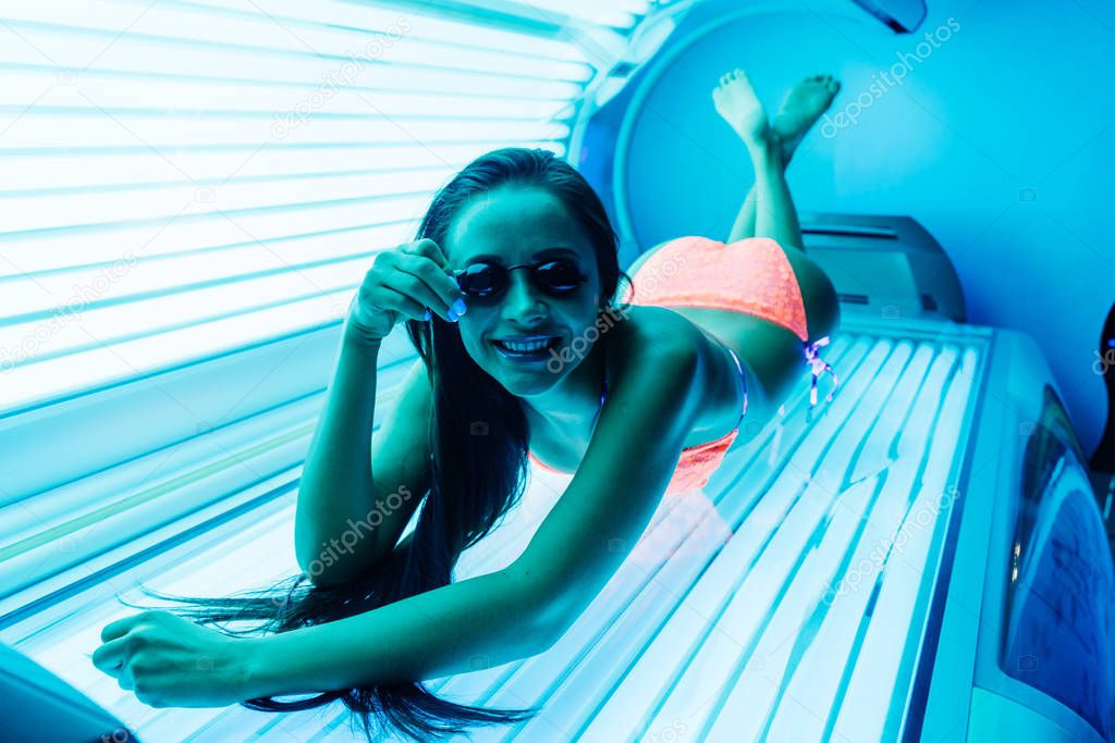 cute young brunette girl in a swimsuit lies in a tanning salon wearing glasses, sunbathing under ultraviolet rays
