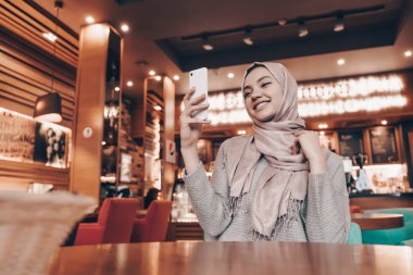 restoranda oturan, ona yiyecek için bekleyen ve smartphone bakarak başörtüsü ile güzel Arap kız gülümseyerek