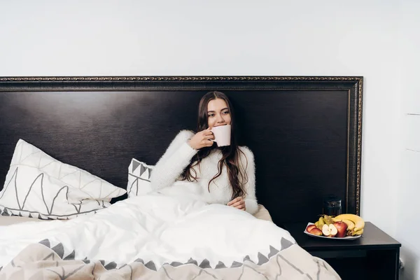 Joven chica de pelo largo en pijama se sienta en la cama temprano en la mañana, bebe café caliente — Foto de Stock