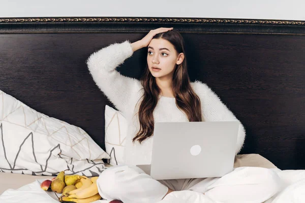Chica joven y confundida freelancer sentado en la cama por la noche y trabajando en su computadora portátil — Foto de Stock