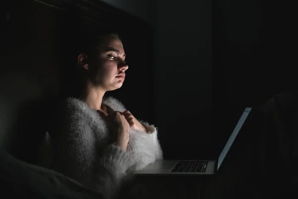 Молодая грустная девушка сидит в постели поздно ночью, смотрит страшный фильм на своем ноутбуке — стоковое фото
