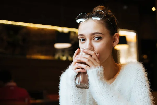 Carino giovane studentessa in un maglione bianco si siede in un caffè, beve un latte macchiato profumato — Foto Stock