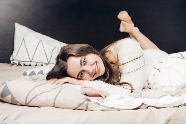 Χαμογελώντας νεαρή κοπέλα σε λευκό πιτζάμες στηρίζεται στο κρεβάτι μετά από την εργασία μιας σκληρής ημέρας — Φωτογραφία Αρχείου