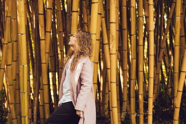 Curly stylish woman in coat posing next to tall bamboo trees in park — Stock Photo, Image