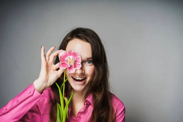 Glad ung kvinna i en rosa skjorta innehar en väldoftande blomma nära ansiktet, ser glad — Stockfoto