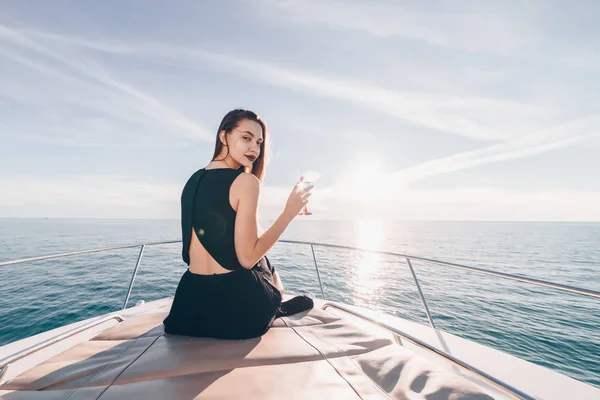 Стильная молодая женщина в черном летнем платье сидит на белой яхте на солнце и пьет шампанское — стоковое фото