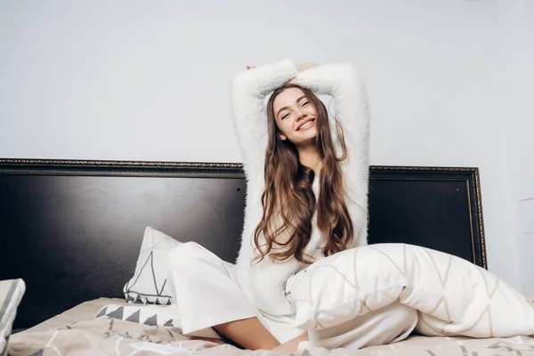 Fröhlich lächelndes Mädchen mit langen Haaren und im Schlafanzug frühmorgens auf dem Bett sitzend, gute Laune — Stockfoto