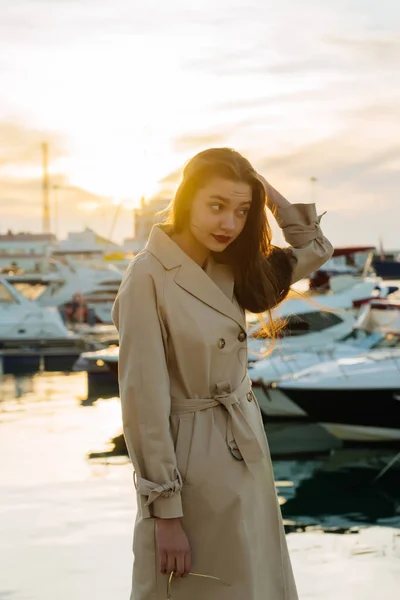 Attractive lovely girl in fashionable beige coat posing in seaport at sunset — Stock Photo, Image