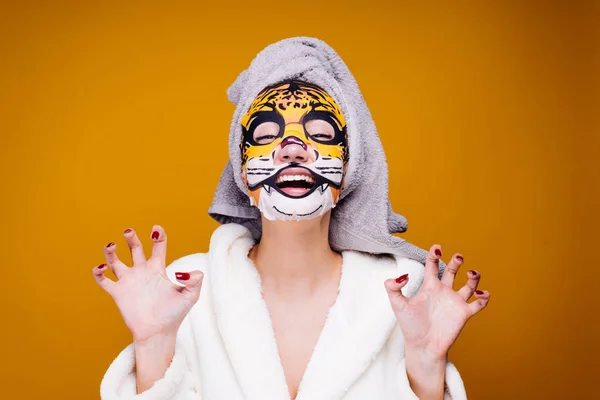 a funny young girl in a white coat and with a towel on her head growls, on her face a moisturizing mask with a leopard face