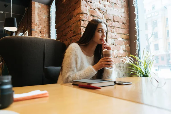 Joven chica de pelo largo pensativa sentada en un café, mirando por la ventana y bebiendo un delicioso café con leche —  Fotos de Stock
