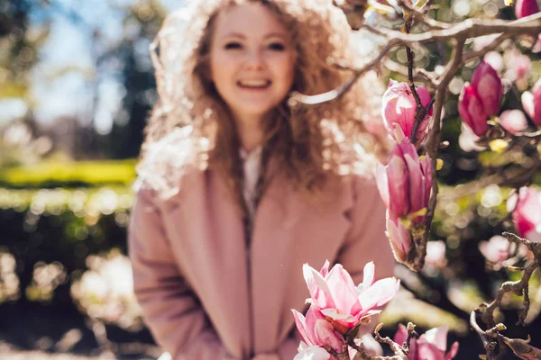 Кучеряве волосся сміється жінка в рожевому пальто позує в парку поруч з запашною магнолією — стокове фото