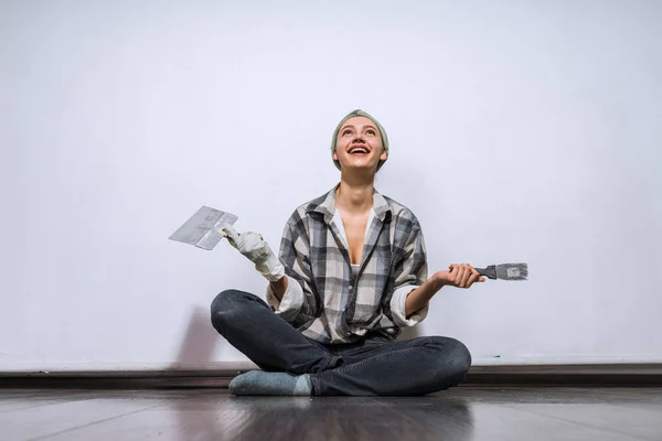 Glimlachend jong meisje in een plaid shirt zittend op de vloer, het vasthouden van een spatel en penseel in handen, het herstellen — Stockfoto