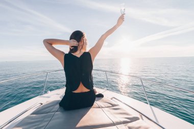 siyah yazlık giysiler içindeki genç bir kız beyaz bir yatta oturur, güneşte şampanya içiyor, bir tatil sahiptir