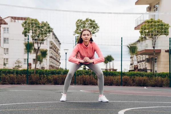 Delgada joven se entrena en un campo de deportes, sentadillas, quiere una figura hermosa — Foto de Stock