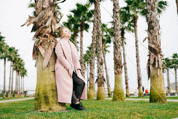 Young woman in long pink coat posing in park next to tall green palm trees — Stock Photo, Image