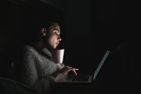 Gece geç saatte yatakta oturuyor ve çay içme karanlık içinde laptop üzerinde çalışan genç kız serbest meslek — Stok fotoğraf