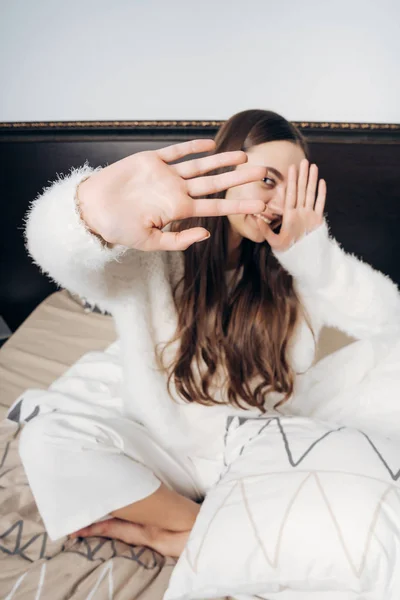 Lustige langhaarige Mädchen im weißen Pyjama sitzt frühmorgens auf dem Bett, schließt sich von der Kamera — Stockfoto
