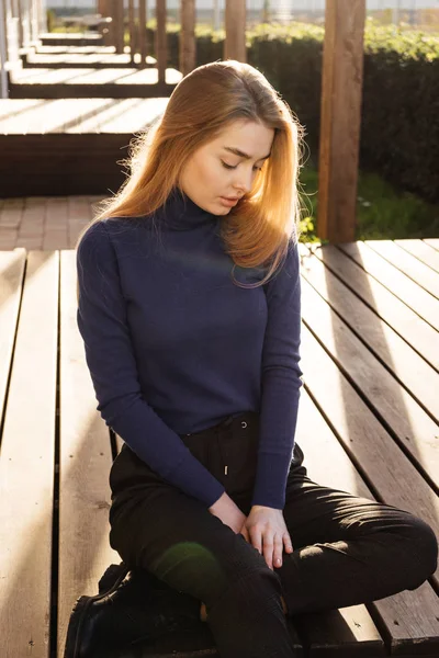 Linda menina loira em uma camisola azul senta-se em um banco no parque, descansando ao sol — Fotografia de Stock