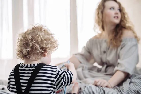 Curly blond little boy in cute clothes sitting on the bed next to her mom — Stock Photo, Image