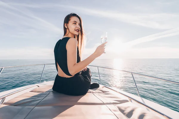 Ein attraktives Mädchen im schwarzen Sommerkleid sitzt auf einer Jacht in der Sonne, trinkt Champagner und genießt die Seereise — Stockfoto