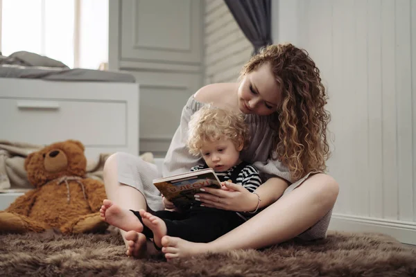 Lockige junge Mutter umarmt ihren kleinen Sohn, liest zusammen ein Buch — Stockfoto