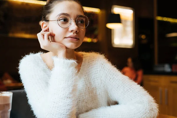 Молодая вдумчивая студентка в очках сидит в кафе после учебы в университете, думая об экзаменах — стоковое фото