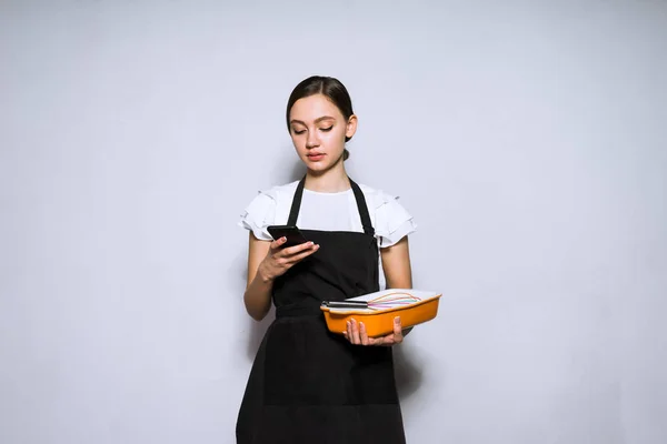 pensive young woman chef chef in black apron cooked cake, looks into her smartphone